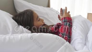 可爱的女孩躺在床上，看着智能手机和社交媒体娱乐。 人们的生活方式和教育。 儿童和儿童